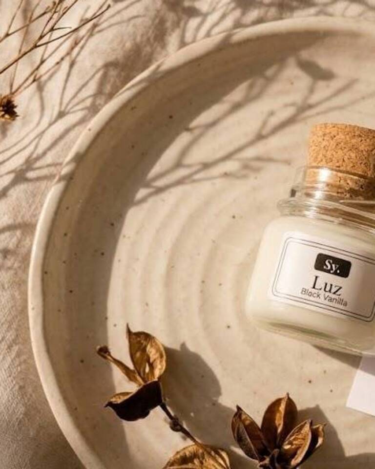 Vela aromática Luz, aroma Black Vanilla, do Studio Yellow sobre prato de cerâmica, com galhos secos ao fundo